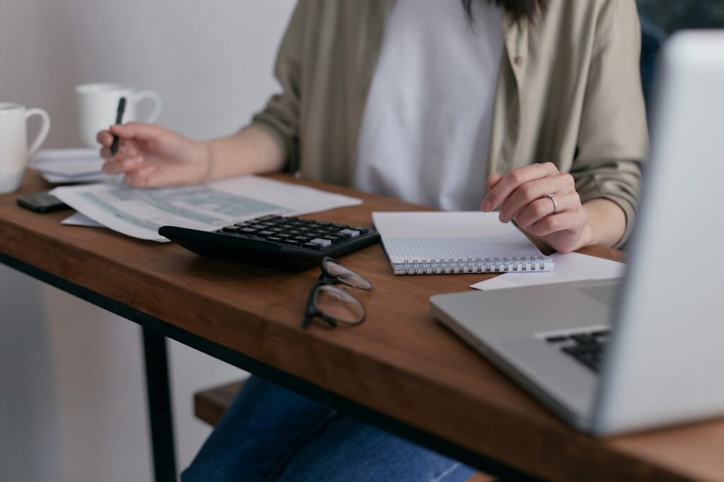 pexels-photo-6963857 A woman manages finances at home, using a laptop and calculator on a wooden desk.